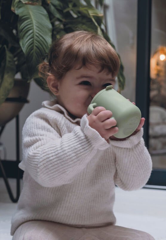 Dziecko w jasnym swetrze pije z zielonego kubka Béaba Sage Green, trzymając go obiema rękami.